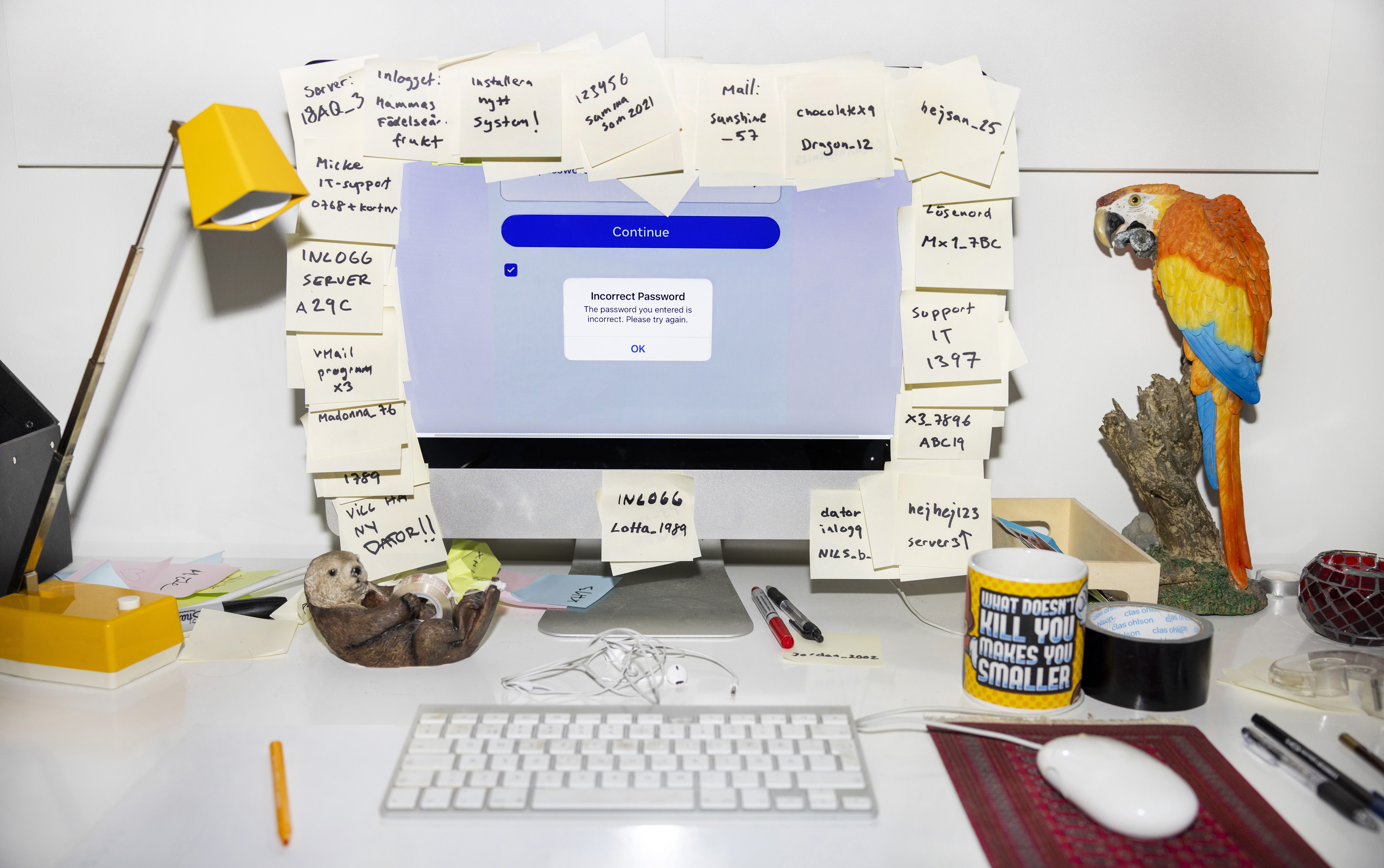 Bildtitel: "Lejonmanen". Bilden visar en vit skrivbordsskiva. I mitten av bilden finns en datorskärm vars ram är täckt av gula post-it-lappar med olika påminnelser och lösenord skrivet i svart. I övrigt finns det vardagliga ting på skrivbordet, som exempelvis en lampa, ett tangentbord, en datormus, en kaffekopp, hörlurar, pennor. Till höger finns även en prydnadspapegoja i rött, gult och blått.