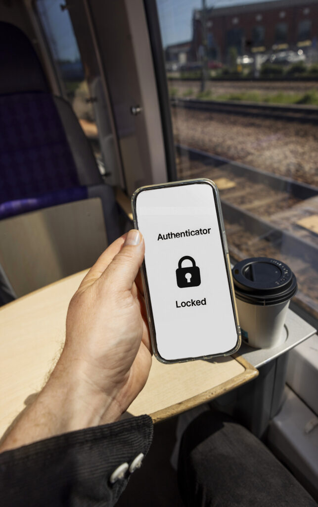 Bildtitel: "Tvåfaktorsautentisering". Bilden visar en hand som håller i en smartphone. På skärmens vita bakgrund syns en hänglåssymbol och texten "Authenticator Locked". Personen befinner sig i en tågvagn och har ett bord och en takeaway-mugg framför sig. Genom fönstret syns tågspår och en byggnad.