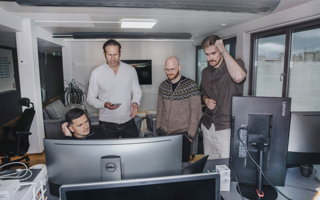 Bildtitel: "It Takes a Village". Bilden visar en kontorsmiljö med en arbetsplats med flera olika datorskärmar. Vid den största datorskärmen sitter en man med bekymrad blick och kliar sig i huvudet. Tre andra män står vid skärmen och ser rådvilla ut. En av dem står med en smartphone i handen.