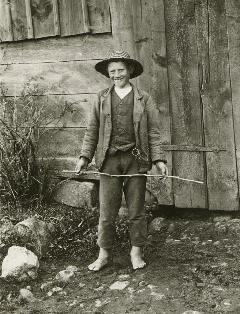 Svartvitt foto. En pojke står utanför en träbyggnad. Han håller i en pinne och ler brett. Han är barfota och klädd i byxor, tröja, väst, en sliten jacka och en flätad hatt.