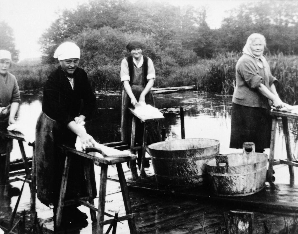 Svartvitt foto. Fyra kvinnor klappar tvätt med klappträn på upphöjda pallar, på en träbrygga. De är klädda i kjolar, uppkavlade yttertröjor och huvuddukar. På bryggan står även två kar i trä. I bakgrunden syns ett vattendrag och växtlighet.