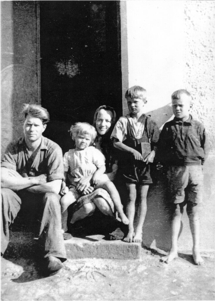 Svartvitt foto. En familj med en pappa, en mamma och tre barn, framför ett murat hus. Föräldrarna sitter på trappan in till huset. Pappan är klädd i långbyxor, uppkavlad arbetsskjorta och träskor. Mamman är klädd i kjol och huvudduk och har ett av barnen i knäet. De två andra barnen står lutade mot väggen. De är barfota och klädda i kortbyxor.